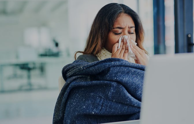 Shot of a young businesswoman blowing her nose with a tissue and covering with a blanket in a modern...