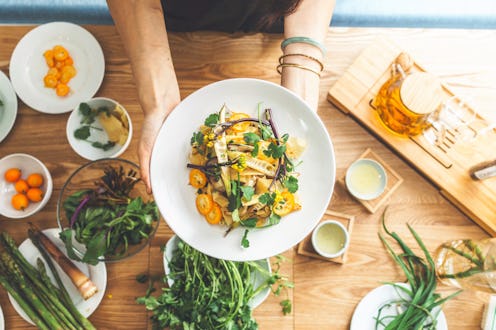 A young woman is holding a vegetarian meal atop a wooden table with wholesome, healthy ingredients i...