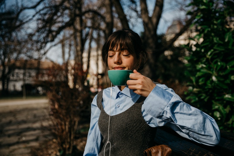 Photo of a beautiful joyful young adult woman sitting outdoors in a park and enjoying her coffee. Sh...