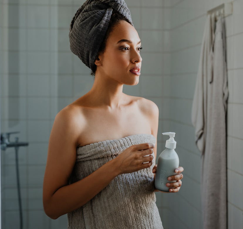 A gorgeous young woman standing in the bathroom after a shower, wrapped in a towel and holding a bot...