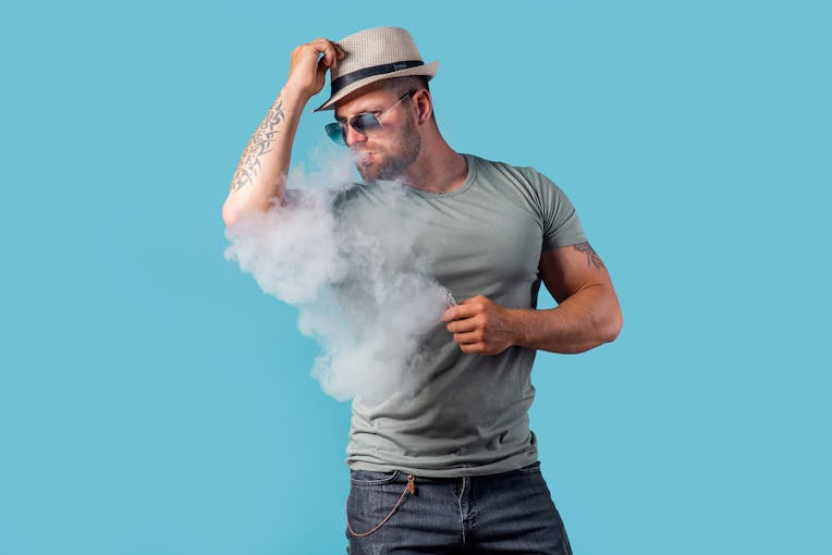 Bearded male in hat and sunglasses smoking a vapor cigarette as an alternative to tobacco. Studio sh...