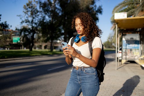 Young attractive Latina woman standing on the city street and waiting for a Taxi to pick her up, usi...