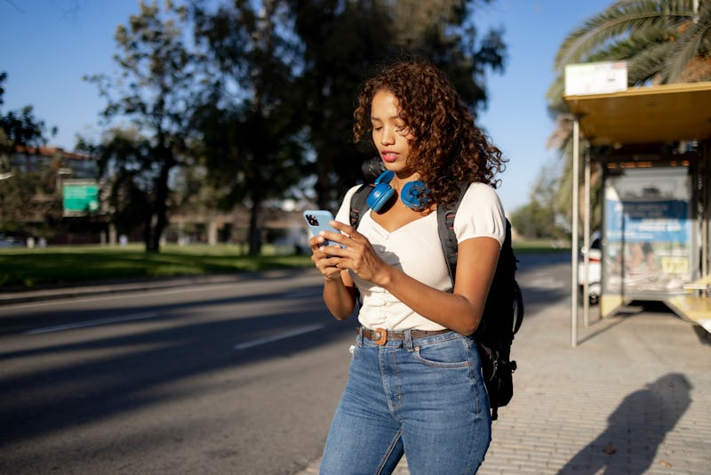 Young attractive Latina woman standing on the city street and waiting for a Taxi to pick her up, usi...