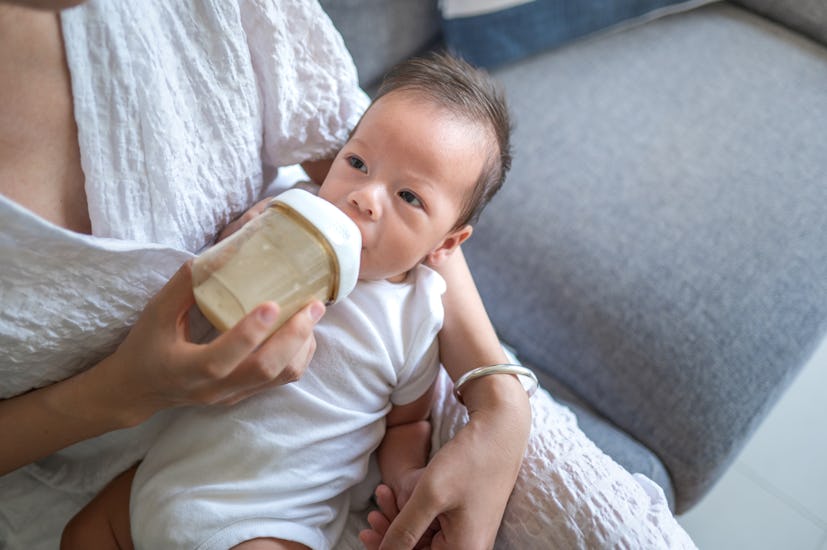 Mother feeding a 40 days old newborn baby boy with a formula milk in a bottle sitting on a sofa in a...