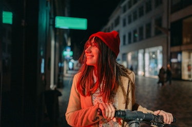 Photo of a young woman riding an e-scooter late at night, in a downtown of a city; illuminated by th...