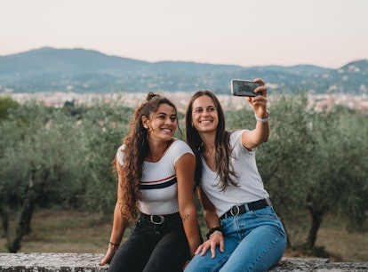 Two friends are taking a selfie together with the countryside in the background. They are sitting on...