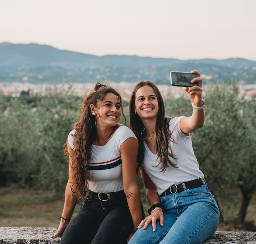 Two friends are taking a selfie together with the countryside in the background. They are sitting on...