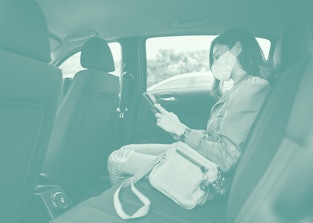 Young woman wearing a protective face mask using a digital tablet while sitting in the backseat of a...