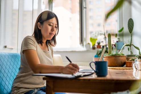 Young Asian woman working with laptop and writing notes at home
