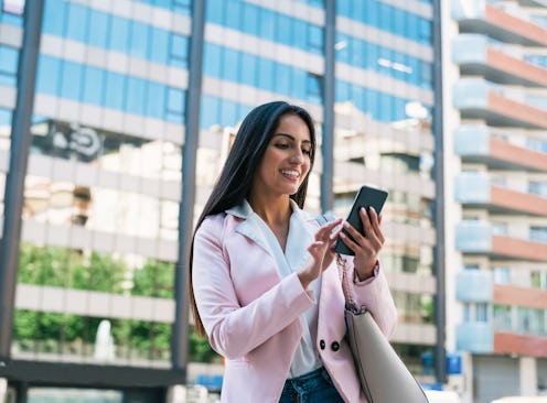 Happy young ethnic female in classy outfit using mobile app on smartphone while standing against con...