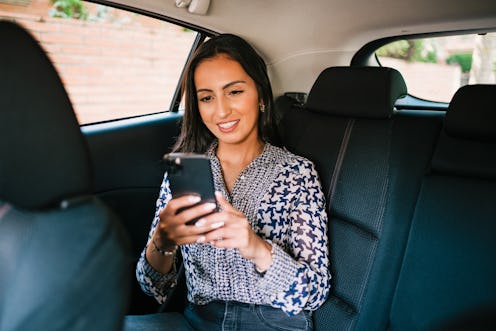Smiling young Hispanic female passenger sitting on backseat of modern car and using mobile phone dur...