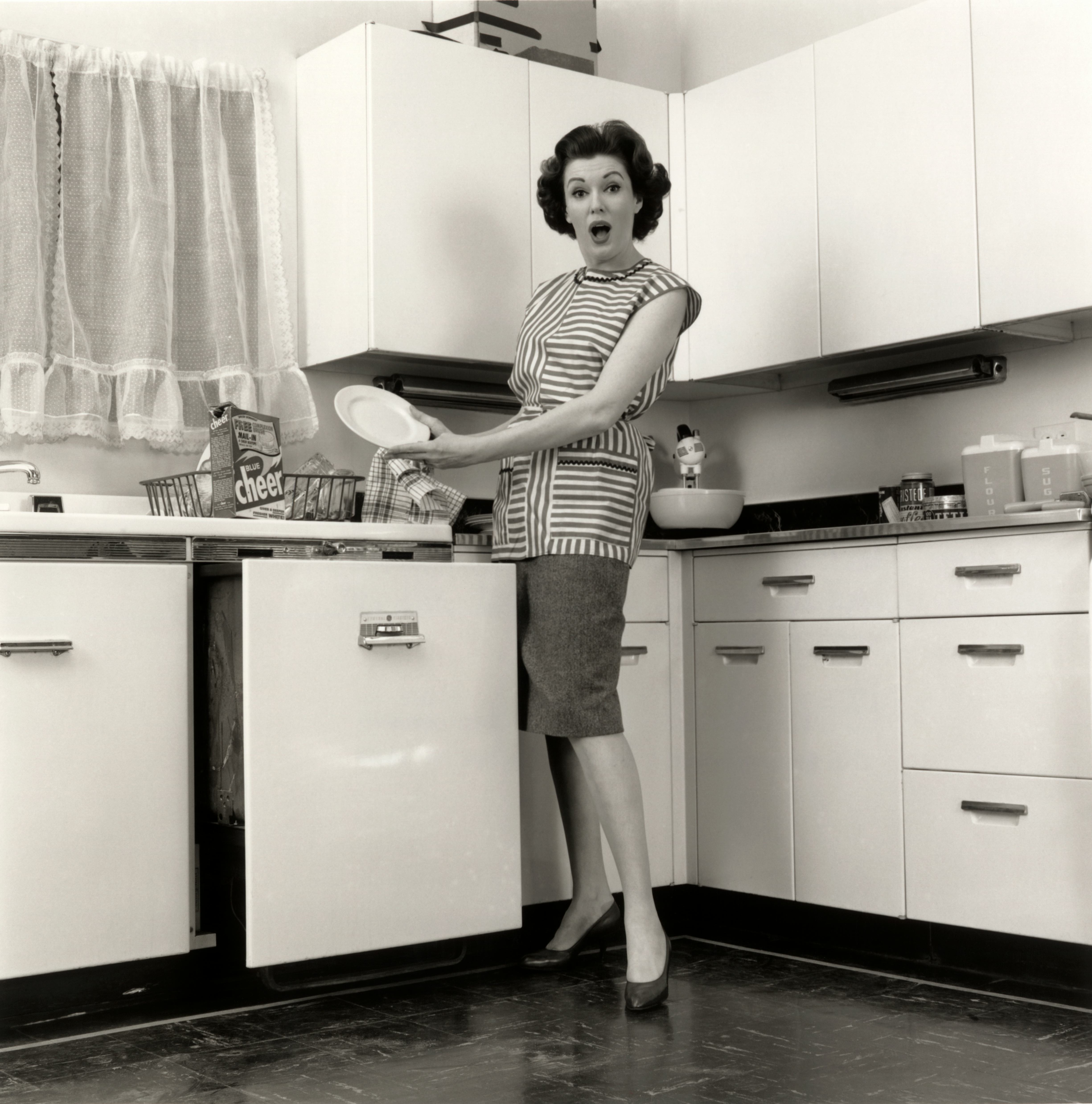 1960s WOMAN HOUSEWIFE WEARING SMOCK PUTTING DISHES IN AUTOMATIC DISHWASHER IN SUBURBAN KITCHEN LOOKI...