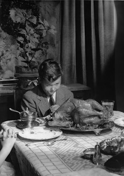 Well dressed boy sitting at table praying while looking at cooked turkey and licking his lips. Circa...