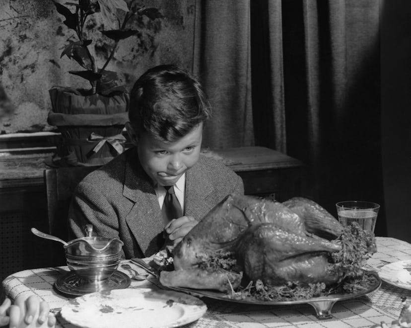 Well dressed boy sitting at table praying while looking at cooked turkey and licking his lips. Circa...