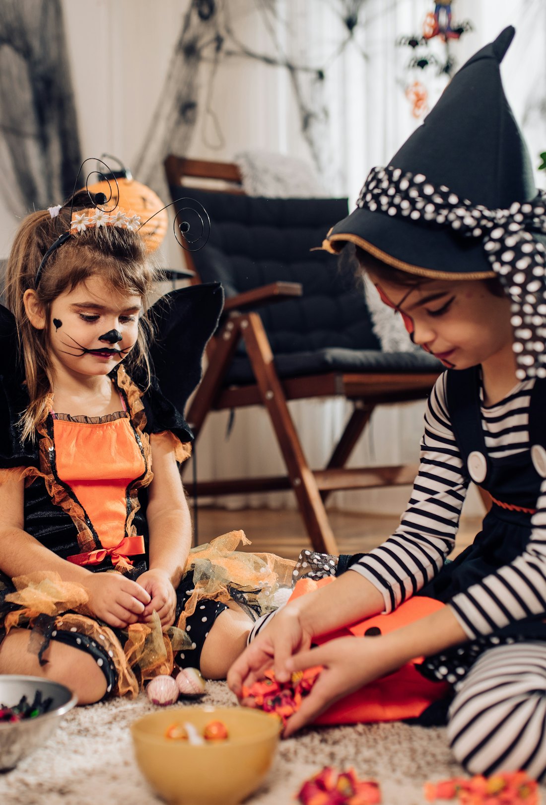 Two cute girls wearing Halloween costumes, playing and sharing candies at home.