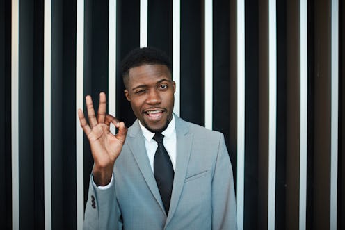 Attractive African American male in gray suit winking and showing OK gesture while standing near str...