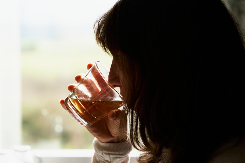 woman drinking whiskey in front the window on a sunny day glass cup between hands