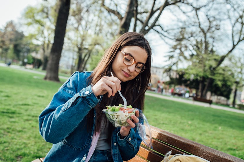 Portrait of a beautiful female student sitting in the park and eating fresh salad.