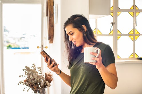 Smiling young woman in pajamas checking her social feeds on her smart phone over a cup of coffee at ...