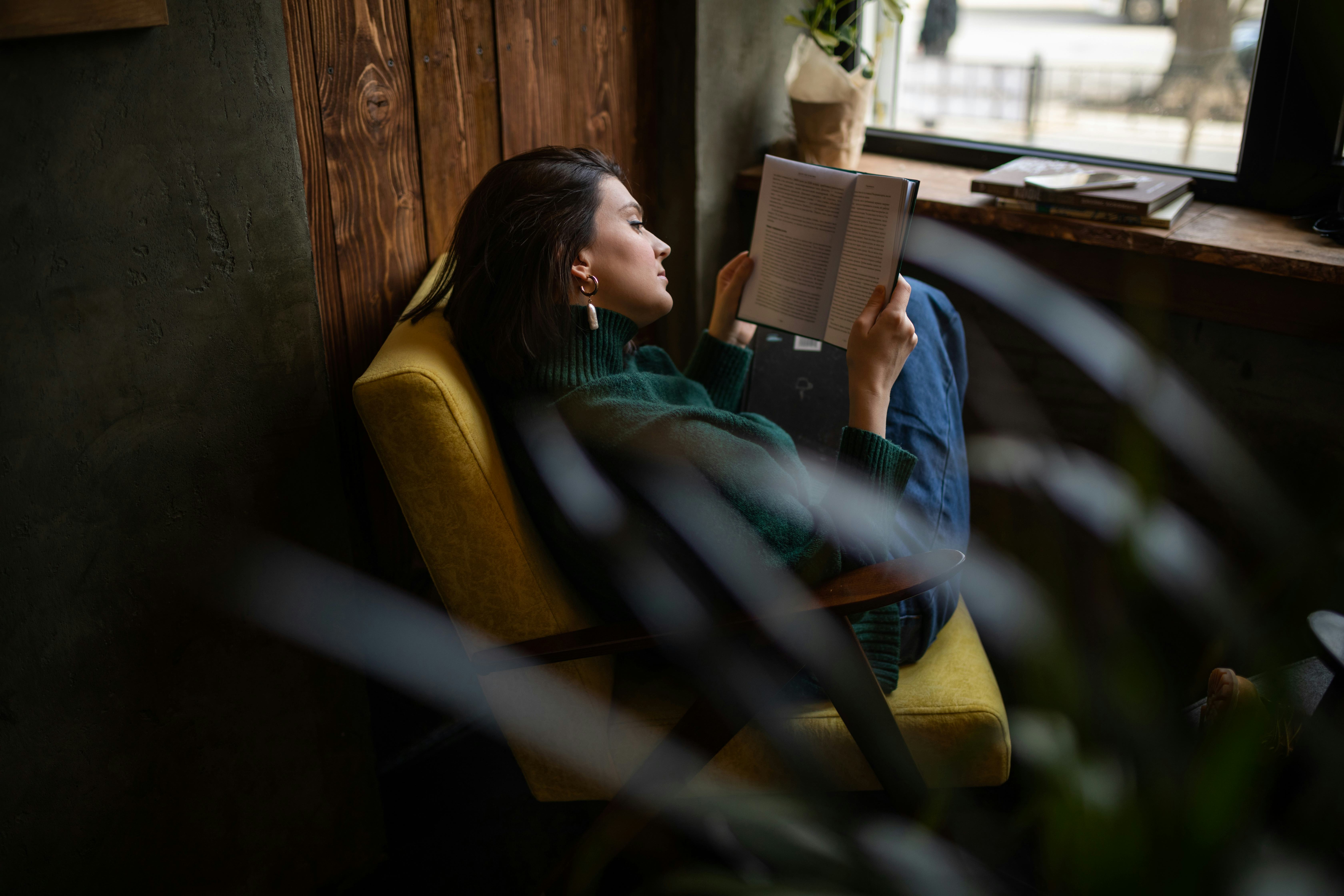Concentrated happy girl sitting on yellow arm chair reading book in modern living room. Young woman ...
