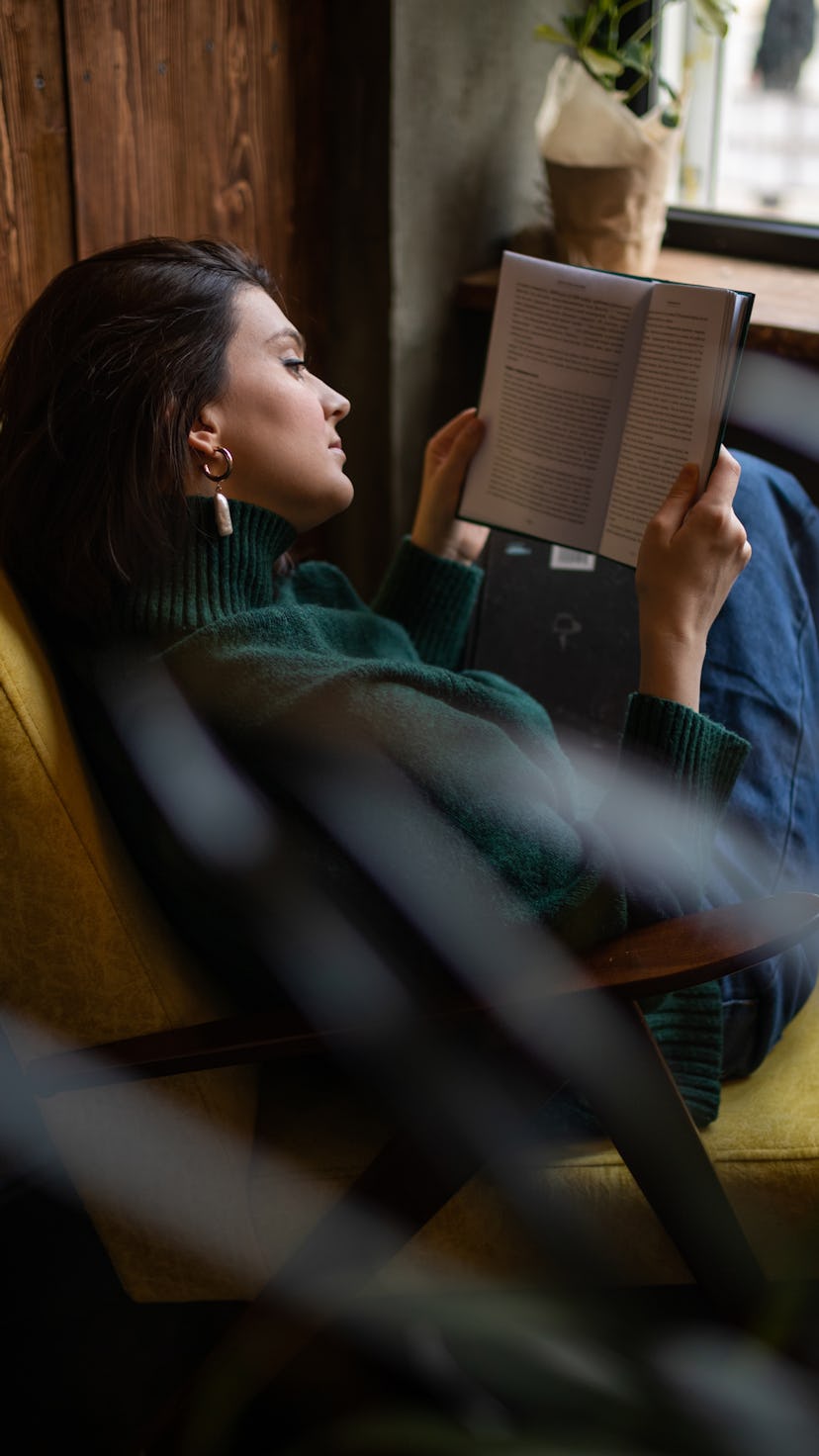 Concentrated happy girl sitting on yellow arm chair reading book in modern living room. Young woman ...