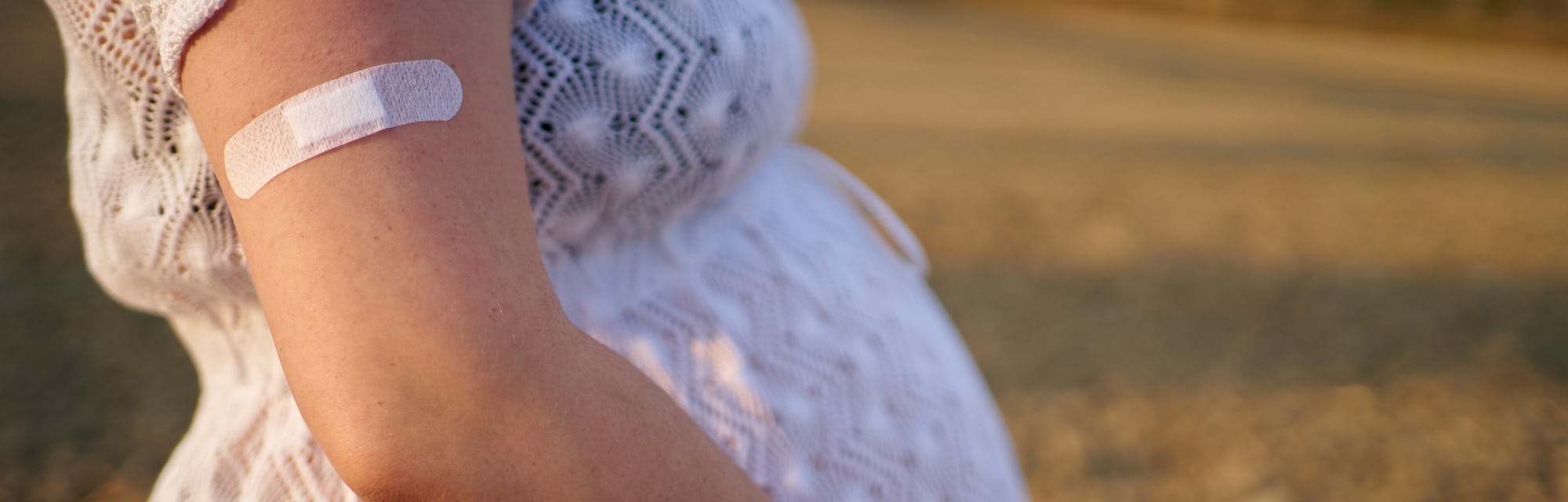 Image of a pregnant person, standing outside with hands on the belly and an adhesive bandage on thei...