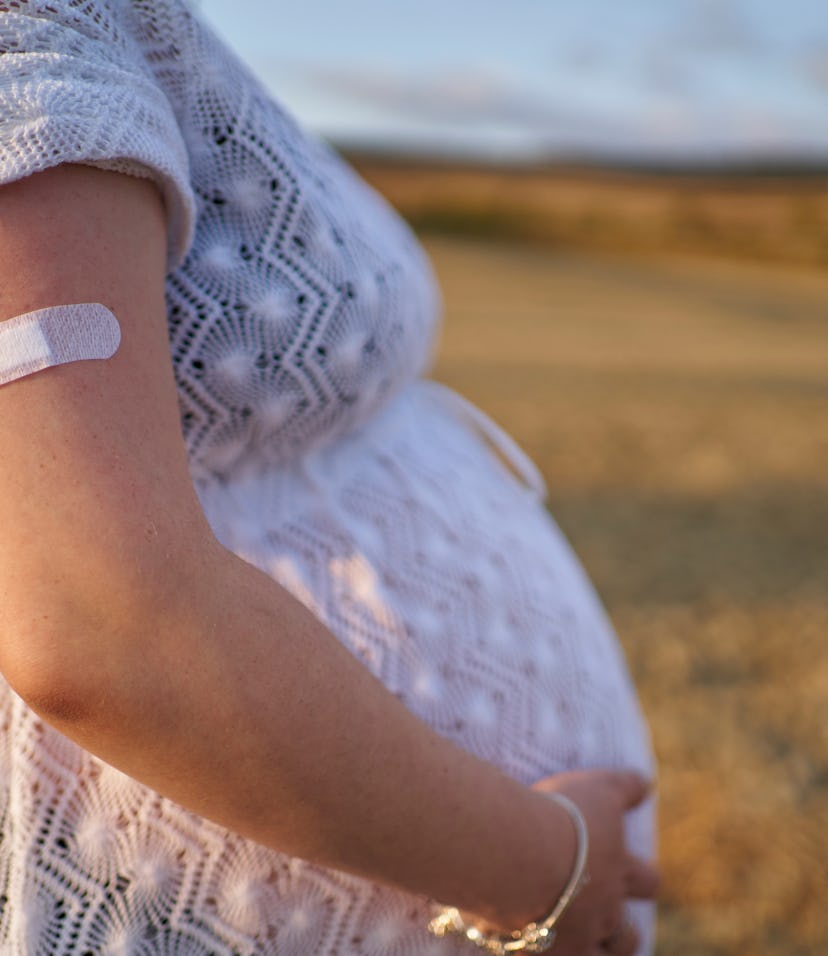 Image of a pregnant person, standing outside with hands on the belly and an adhesive bandage on thei...