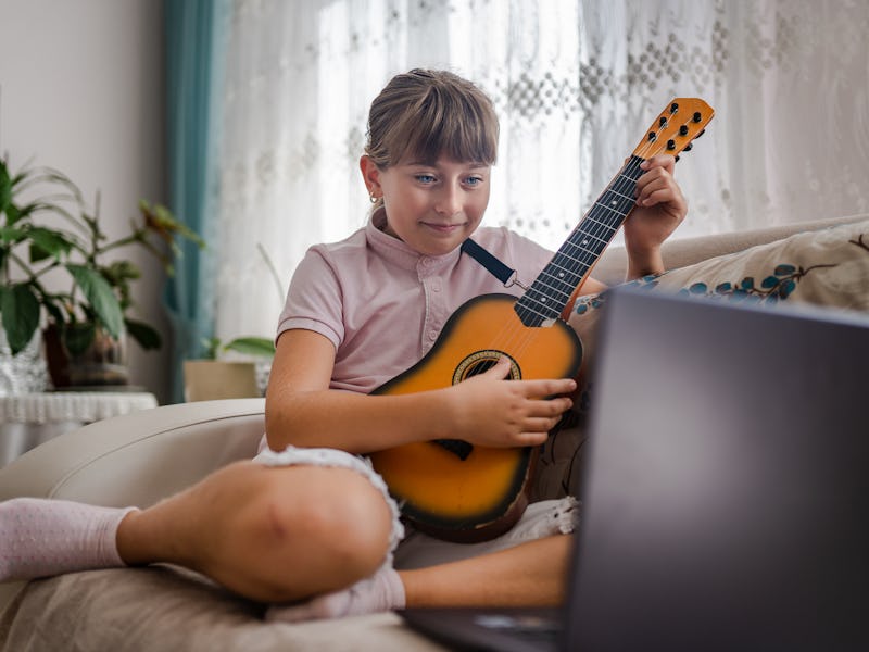Little girl taking guitar lesson online and playing guitar against laptop