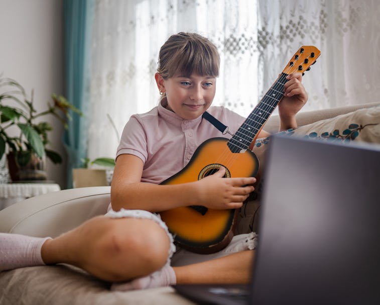 Little girl taking guitar lesson online and playing guitar against laptop