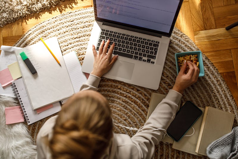 Directly above shot of diligent teenager lying on the floor, snacking and using laptop while doing h...