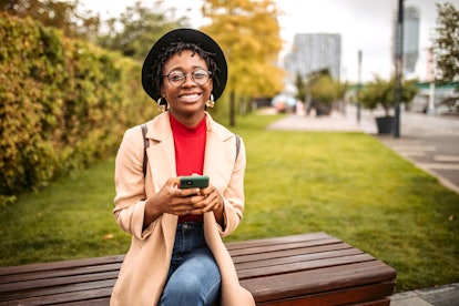 Young, cute, smiling, black woman sitting on the bench in public park, enjoying autumn day, using sm...