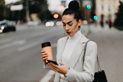 Young elegant woman standing on the sidewalk in the city and waiting Uber taxi. Holding cup of coff...