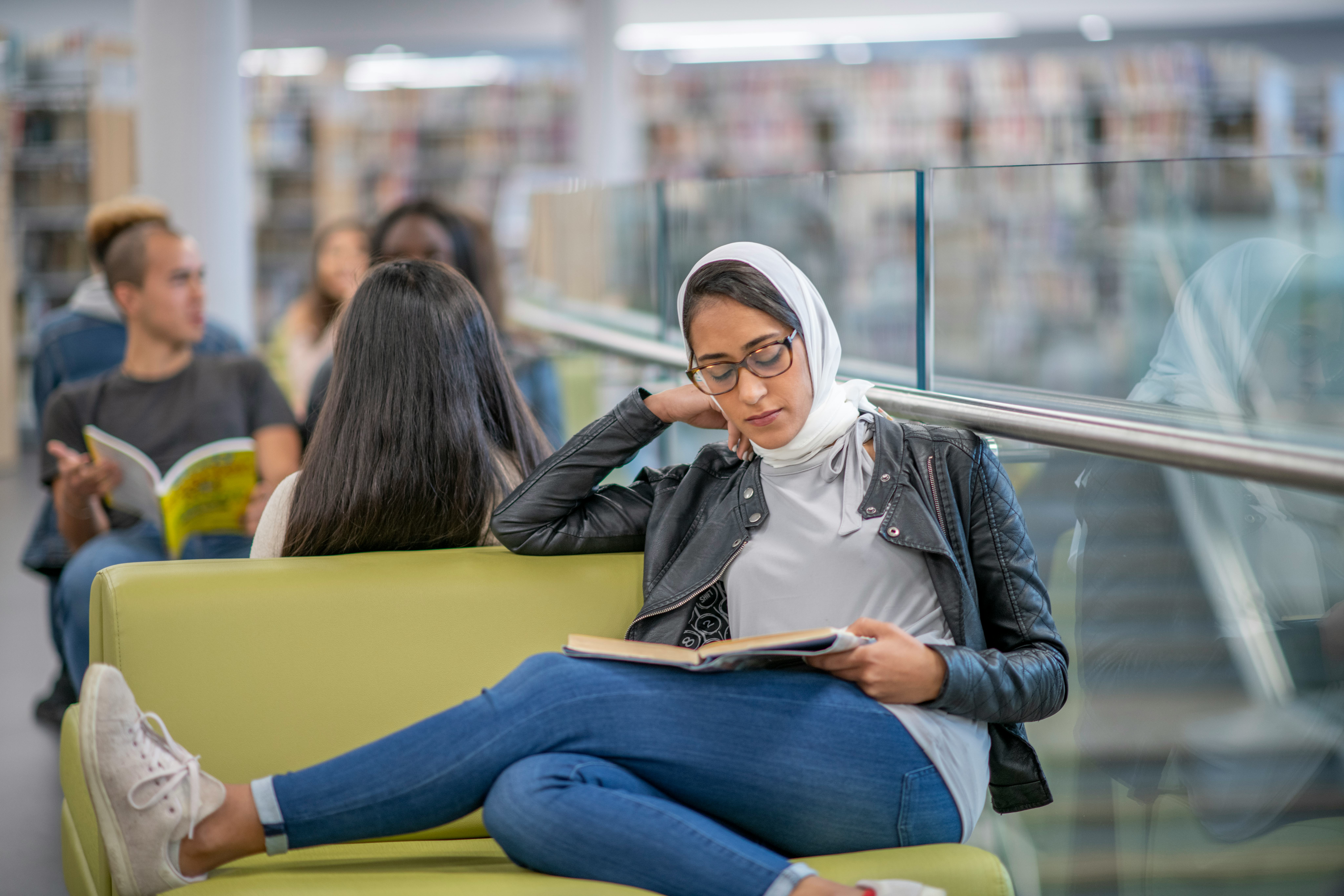 A Middle Eastern woman wearing a hijab sits at a chair beside a railing and studies.