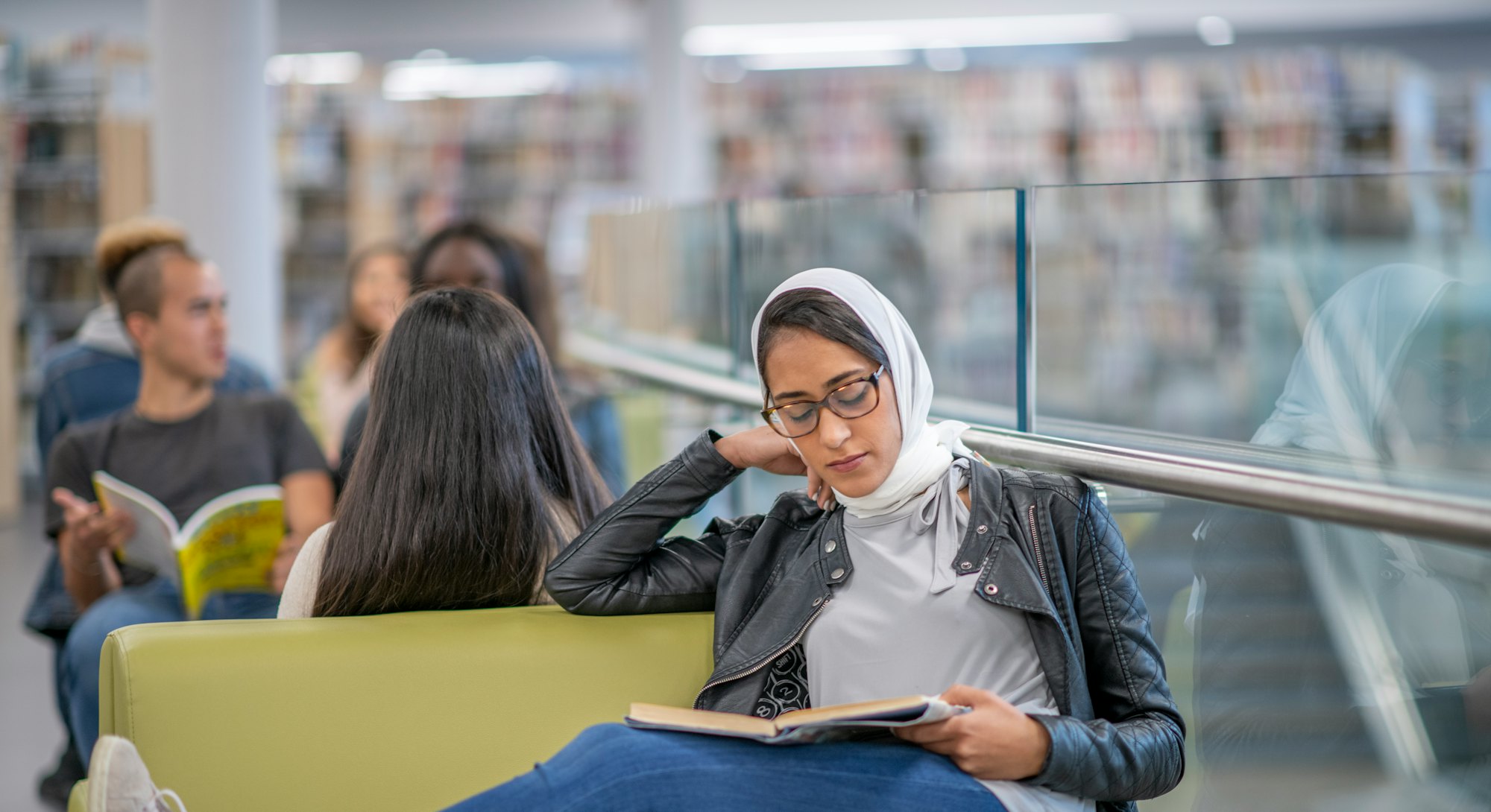 A Middle Eastern woman wearing a hijab sits at a chair beside a railing and studies.