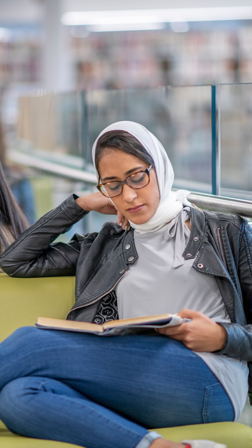 A Middle Eastern woman wearing a hijab sits at a chair beside a railing and studies.