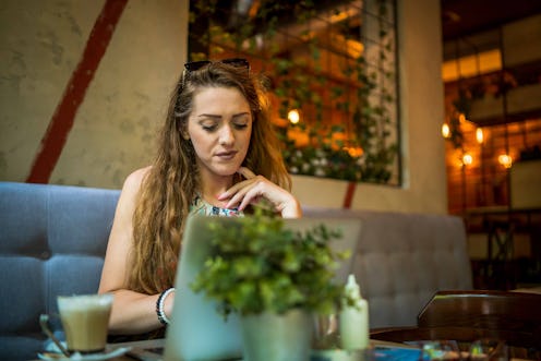 Young woman sitting in coffee shop at wooden table, drinking coffee. On table is laptop. Girl browsi...