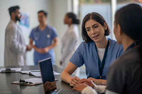 Two female medical professionals sit at a desk with a laptop open in front of them as they review a ...