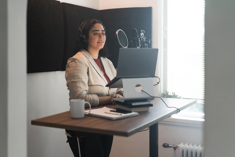 Woman recording a podcast from her home studio. She is standing by her desk, on it is her laptop and...