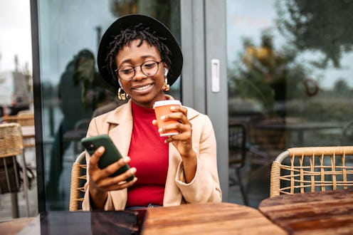 Young fashionable smiling black business woman sitting in outdoor cafe on rainy autumn day, using sm...