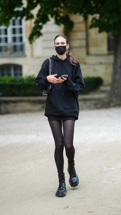 A model wears a black oversized hoodie sweaters, black tights, a black skirt, black shiny leather la...