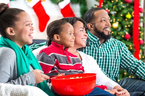 Image of four people on a couch with a bowl of popcorn. A Christmas tree and holiday stockings can b...