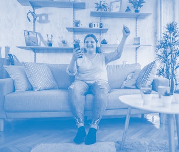 Excited young overweight woman holding mobile phone singing and dancing at home.