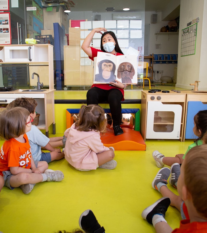 A teachers wearing a protective face mask reads a book to children while making dramatic faces on Ju...