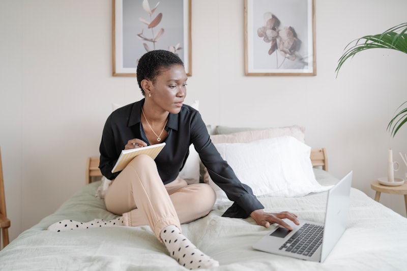 Black woman taking notes in a notebook while using a computer laptop on a bed at home. Technology, b...