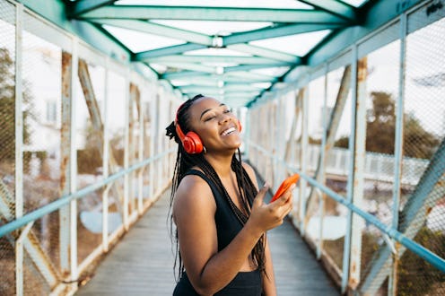 Portrait of a young smiling woman, with braided hair and sportswear, using a red smartphone and head...
