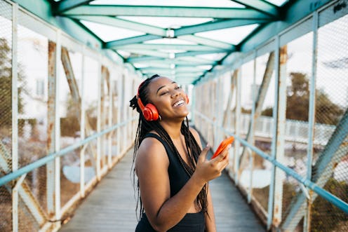 Portrait of a young smiling woman, with braided hair and sportswear, using a red smartphone and head...