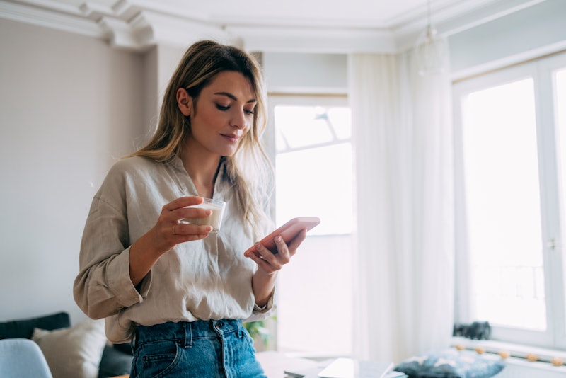 Young smiling female holding cup of coffee and typing text message on her smartphone in the morning