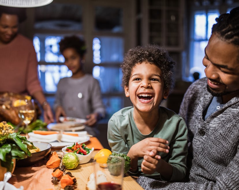 Cheerful African American boy laughing while sitting in his father's lap during Thanksgiving day at ...