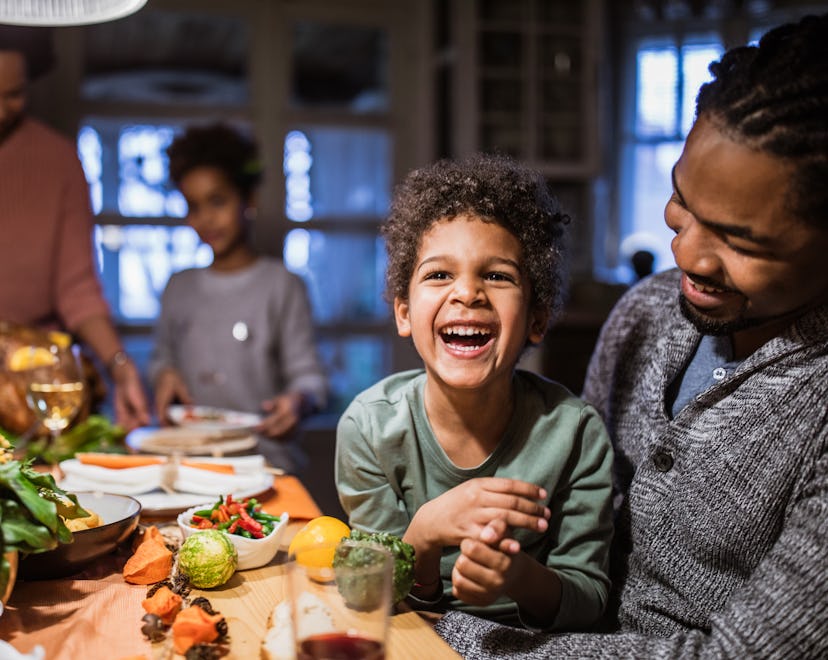 Cheerful African American boy laughing while sitting in his father's lap during Thanksgiving day at ...