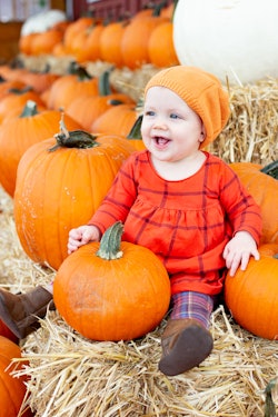 Image of a baby sitting on a hay bale, surrounded by pumpkins. Baby is wearing an orange dress and h...